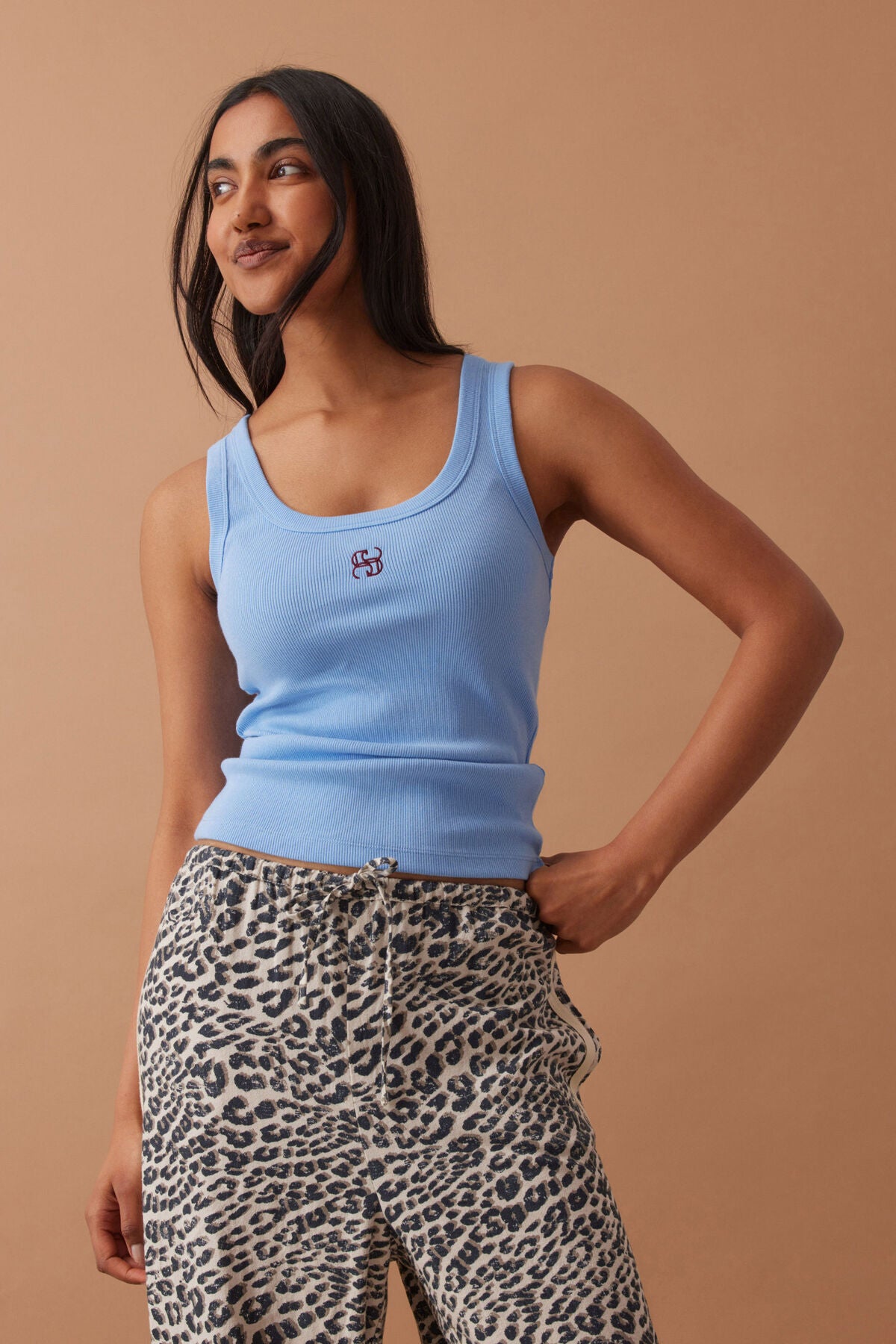 Woman wearing a light blue tank top with a logo and patterned pants on a beige background